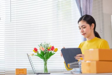 Bir iş kadını, siparişleri göndermeden önce evindeki ürünleri dağıtmak ve siparişleri kontrol etmek için internet üzerinden satış işine yatırım yapar..