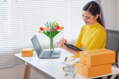 Bir iş kadını, siparişleri göndermeden önce evindeki ürünleri dağıtmak ve siparişleri kontrol etmek için internet üzerinden satış işine yatırım yapar..