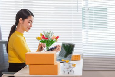 Bir iş kadını, siparişleri göndermeden önce evindeki ürünleri dağıtmak ve siparişleri kontrol etmek için internet üzerinden satış işine yatırım yapar..