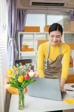 Bir iş kadını, siparişleri göndermeden önce evindeki ürünleri dağıtmak ve siparişleri kontrol etmek için internet üzerinden satış işine yatırım yapar..