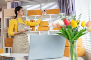 Bir iş kadını, siparişleri göndermeden önce evindeki ürünleri dağıtmak ve siparişleri kontrol etmek için internet üzerinden satış işine yatırım yapar..