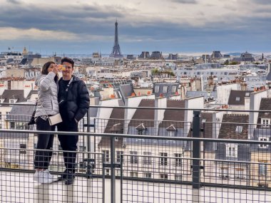 Paris Fransa - 04.06.2022: Paris 'in çatısında genç bir çift, arka planda Eyfel kulesi ve güzel bir genital şehir manzaralı cep telefonuyla selfie çekiyor. Kopyasıyla bulutlu gökyüzü 