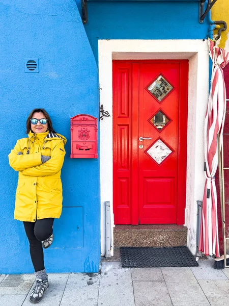 Happy traveler woman tourist with yellow raincoat and sun glasses posing at the entrance of a blue house with red door and postbox on Burano island in Venetian lagoon. Winter in Venice, Italy concept.