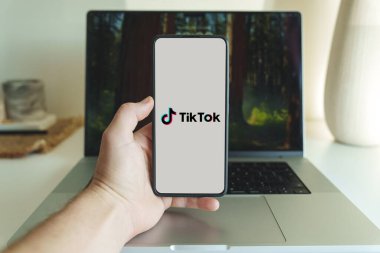 Londra, İngiltere - 19 Ekim 2024: Cep telefonu ekranında TikTok uygulaması logosu. Bilgisayarın arka planında Tiktok uygulaması olan kişi
