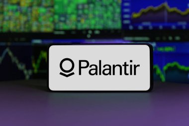 Palantir logosu, arka planda borsa çizelgeleri olan bir akıllı telefon ekranında sergileniyor. 2 Ocak 2025