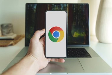 Google Chrome logosunu arka planda dizüstü bilgisayarla gösteren akıllı telefonu tutan bir el. 3 Ocak 2025