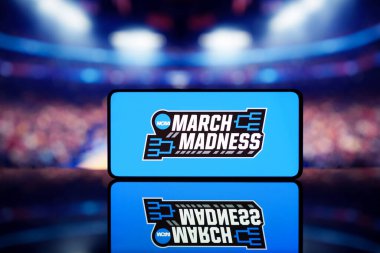 Arka planda bulanık basketbol sahası olan March Madness logosunu gösteren bir akıllı telefon. 13 Ocak 2025