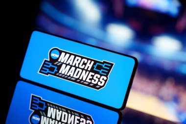 Arka planda bulanık basketbol sahası olan March Madness logosunu gösteren bir akıllı telefon. 13 Ocak 2025