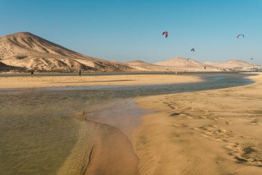 Fuerteventura 'daki kumsalda dalgalı bir akarsu okyanusa akıyor. Arka planda dağlar var ve mavi gökyüzünün altında birkaç uçurtmacı görülebiliyor. Yüksek kalite fotoğraf