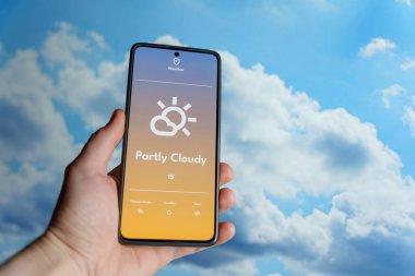 Bir hava durumu uygulamasını parçalı bulutlu bir hava tahminiyle gösteren akıllı bir telefon tutuyorum. Arka planda dağınık bulutlar olan mavi gökyüzü. Hava teknolojisi ve hava tahminleri. Yüksek kalite fotoğraf