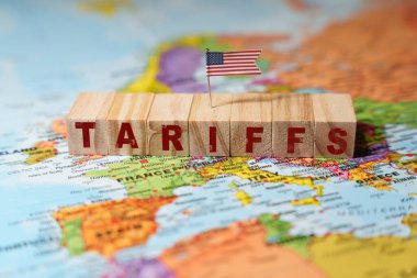 Tahta bloklar üzerinde ABD bayrağı olan TARIFFS yazılıyor, Avrupa haritasında küresel ticaret politikalarını ve ekonomik etkiyi sembolize ediyor. Yüksek kalite fotoğraf
