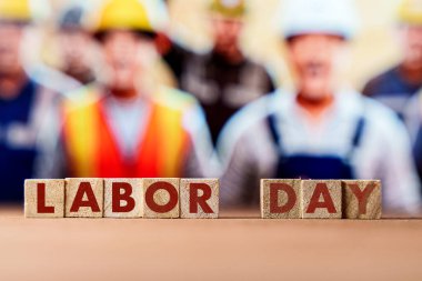 Tahta bloklar LABOR Günü 'nü bulanık inşaat işçilerinin güvenlik kaskı ve yelek giydiği bir yüzeye koyuyor. İşçi memnuniyeti, iş hakları ve istihdam kutlamaları. 