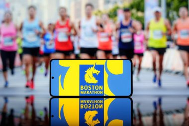 Arka planda bulanık koşucuların olduğu Boston Maratonu logosunu gösteren akıllı telefon. Ünlü koşu etkinliği ve spor yarışması konsepti. 17 Mart 2025