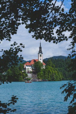 Bled Gölü 'ndeki İkonik Varsayım Kilisesi, ağaçların arasına kurulmuş ve yeşil ağaç yapraklarıyla çerçevelenmiş. Yüksek kalite fotoğraf