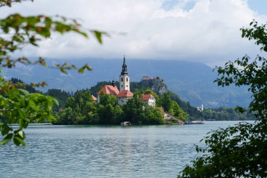 Bled Gölü 'ndeki İkonik Varsayım Kilisesi, ağaçların arasına kurulmuş ve yeşil ağaç yapraklarıyla çerçevelenmiş. Yüksek kalite fotoğraf