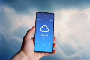 Bulutlu bir hava tahminine sahip bir hava durumu uygulamasını gösteren akıllı bir telefon tutuyorum. Bulanık arka planda gökyüzünü dolduran yumuşak bulutlar. Hava teknolojisi ve hava tahminleri. Yüksek kalite fotoğraf
