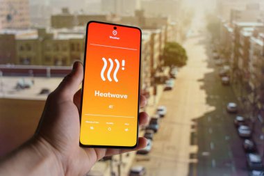 Sıcak hava dalgası uyarısı olan bir hava durumu uygulamasını gösteren akıllı bir telefon. Arka planda aşırı ısı altında bulanık şehir manzarası. Yüksek sıcaklık ve iklim değişikliği kavramı. Yüksek kalite fotoğraf