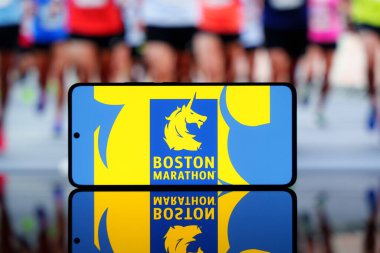 Arka planda bulanık koşucuların olduğu Boston Maratonu logosunu gösteren akıllı telefon. Ünlü koşu etkinliği ve spor yarışması konsepti. 17 Mart 2025