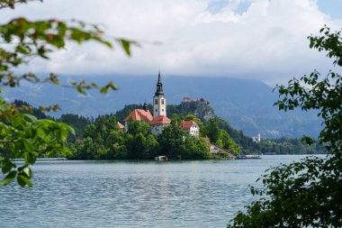 Bled Gölü 'ndeki İkonik Varsayım Kilisesi, ağaçların arasına kurulmuş ve yeşil ağaç yapraklarıyla çerçevelenmiş. Yüksek kalite fotoğraf
