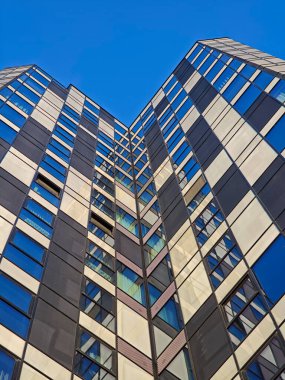 Yerden görülen modern gökdelen cephesinde, koyu renk panellerden oluşan tekrarlayan geometrik bir dama tahtası deseni ve berrak mavi gökyüzüne uzanan yansıtıcı mavi cam pencereler yer alıyor. Yüksek kalite fotoğraf