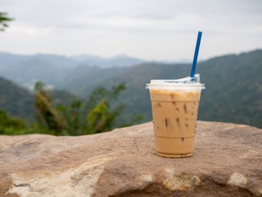A glass of iced coffee is placed on a rock against a background of mountains and sky.