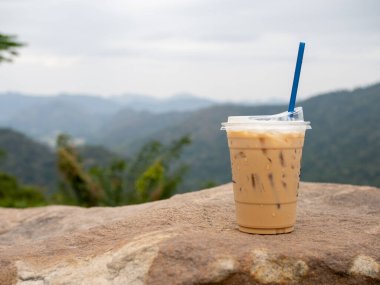 A glass of iced coffee is placed on a rock against a background of mountains and sky.