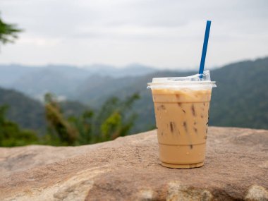 A glass of iced coffee is placed on a rock against a background of mountains and sky.
