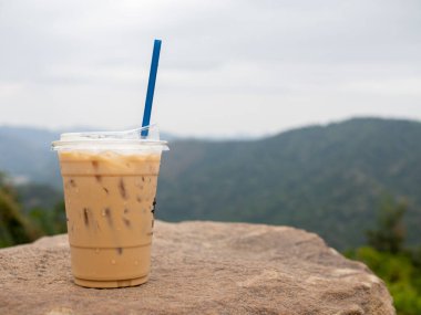 A glass of iced coffee is placed on a rock against a background of mountains and sky.