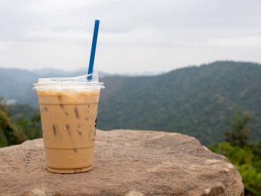 A glass of iced coffee is placed on a rock against a background of mountains and sky.