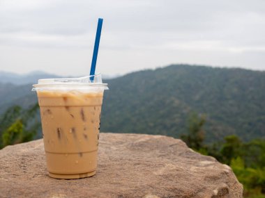 A glass of iced coffee is placed on a rock against a background of mountains and sky.