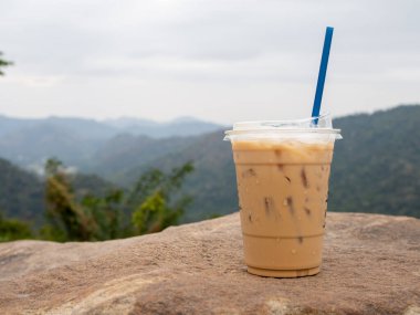 A glass of iced coffee is placed on a rock against a background of mountains and sky.