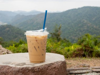 A glass of iced coffee is placed on a rock against a background of mountains and sky.
