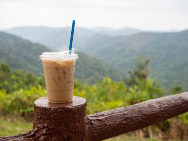 A glass of iced coffee is placed on the fence against a backdrop of mountains and sky.