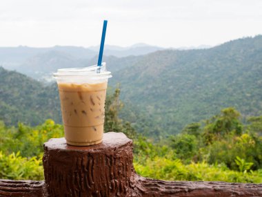 A glass of iced coffee is placed on the fence against a backdrop of mountains and sky.