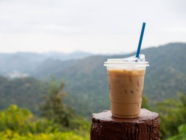 A glass of iced coffee is placed on the fence against a backdrop of mountains and sky.
