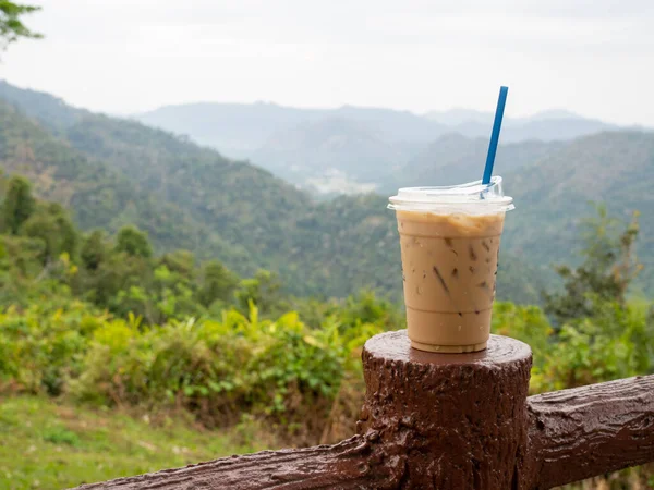 A glass of iced coffee is placed on the fence against a backdrop of mountains and sky.