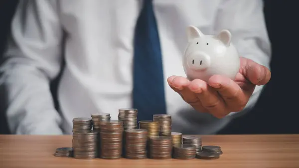 Human holds a piggy bank and there are coins lined up on the wooden floor. Concepts of finance, savings, investment, and setting goals for fixed deposits with banks.