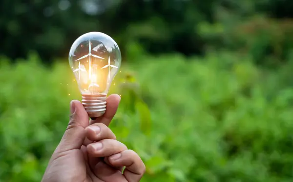 Concept of renewable energy, environmental protection, and sustainable renewable energy sources. Person\'s hand holding a wind turbine in a light bulb on a natural green background. Clean energy.