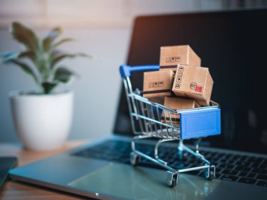 Dizüstü bilgisayara yerleştirilen alışveriş arabası ve ürün kutuları çevrimiçi alışveriş kavramı, web sitesi, e-ticaret, pazar platformu, teknoloji, ulaşım, lojistik ve çevrimiçi ödeme kavramını temsil etmektedir..