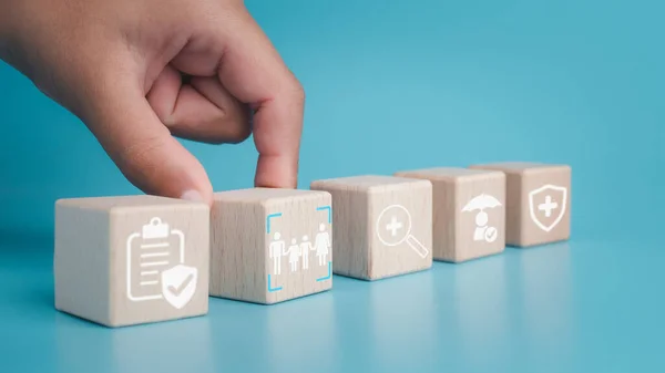 Health insurance and healthcare concept, human hand holds wooden block with icons about health insurance and healthcare access, retirement planning on blue background.