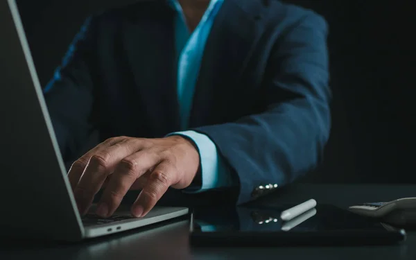Businessman in black suit working on laptop computer and tablet, Hand typing on keyboard on table at office with dark background, Online working, Close up, Copy space.