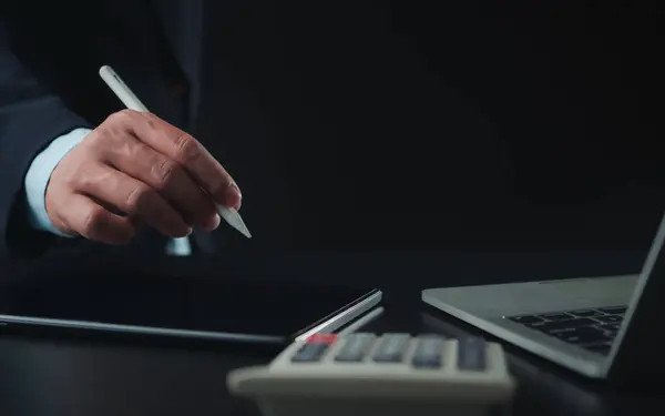 Businessman in black suit working on laptop computer and tablet and calculator, Hand holding stylus pen touch on tablet at office with dark background, Online working, Close up, Copy space.