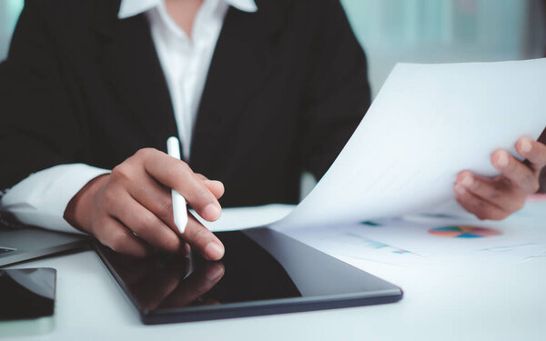 Businesswoman in black suit working on laptop computer, paper documents, tablet, and calculator, Hands touch on tablet at office with dark background, Online working, Close up, Copy space.