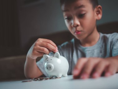 Asyalı çocuk kumbarada para biriktiriyor, bir şeyler düşünmeyi öğreniyor, çocuk gelecek eğitim için para biriktiriyor. Para, finans, sigorta ve insan konsepti.