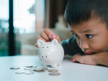 Asyalı çocuk kumbarada para biriktiriyor, bir şeyler düşünmeyi öğreniyor, çocuk gelecek eğitim için para biriktiriyor. Para, finans, sigorta ve insan konsepti.