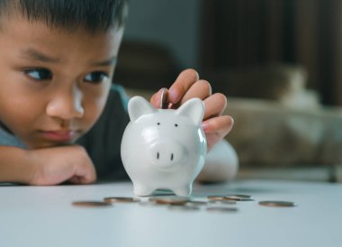 Asyalı çocuk kumbarada para biriktiriyor, bir şeyler düşünmeyi öğreniyor, çocuk gelecek eğitim için para biriktiriyor. Para, finans, sigorta ve insan konsepti.