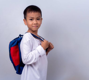 Beyaz giyinmiş, beyaz çantalı, beyaz arka planda duran Asyalı sevimli çocuk mutlu ve mutlu görünüyor..