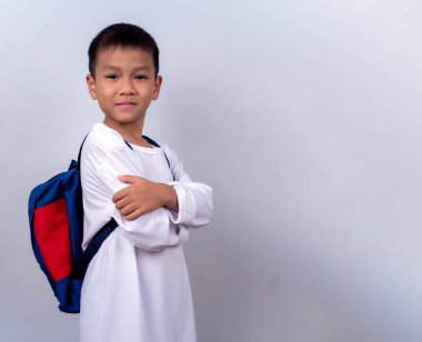 Beyaz giyinmiş, beyaz çantalı, beyaz arka planda duran Asyalı sevimli çocuk mutlu ve mutlu görünüyor..