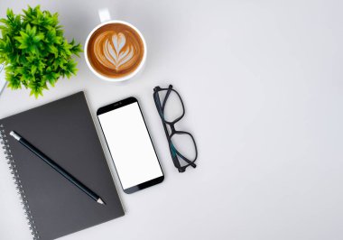 Boş ekran akıllı telefonu, kahve fincanı, defter, kalem, gözlük ile modern çalışma alanının üst görüntüsü. Klavye ve aksesuarlarla ilgili ofis durumu beyaza izole edildi. Uzayı kopyala, düzleştir.