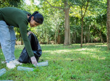 Gönüllü bir kadın parktaki bir çevre temizleme etkinliğine katılıyor. Eldiven takan ve büyük siyah çantalar tutan bir kadın. Parkta çöp ve plastik şişe toplayan bir kadın..